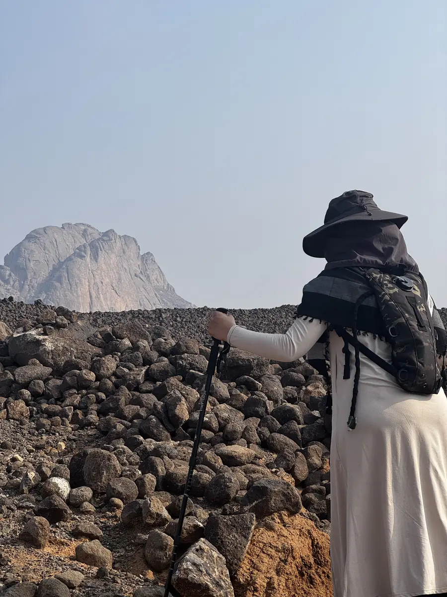 A sister with a trekking pole walking over volcanic rock towards a pale peak